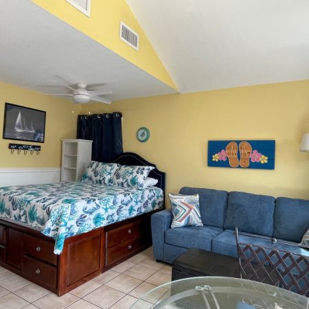 Bright studio room with a bed, blue sofa, tropical decor, tile floor, and yellow walls.