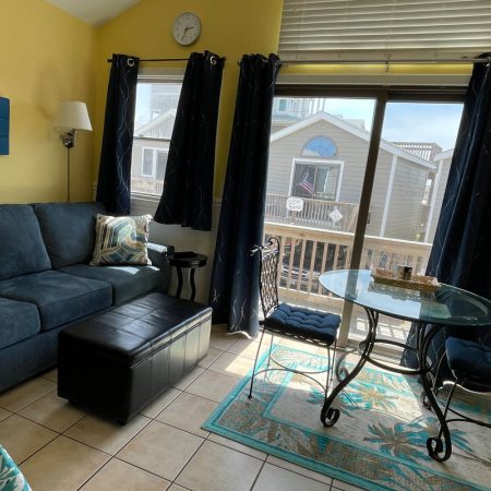 Living room with a blue sofa, round glass table, two chairs, and sunlight coming through large windows and a sliding door.