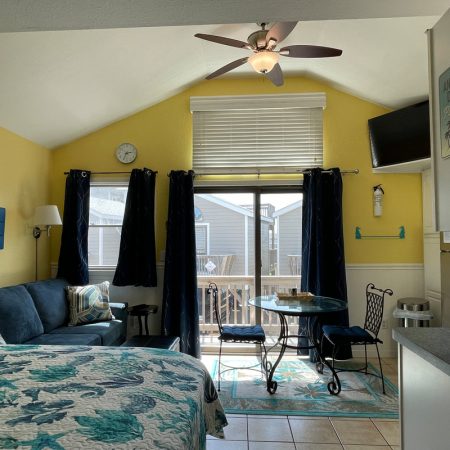 Sunny living area with yellow walls, blue sofa, glass table, and sliding doors leading to a deck outside.