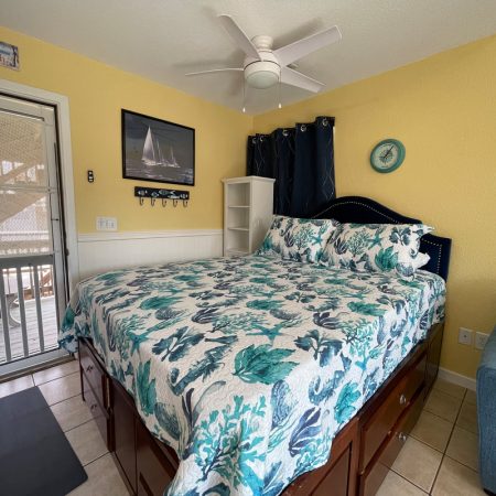 Coastal-themed bedroom with a bed, blue floral bedding, ceiling fan, shelf, and yellow walls near a door with screen.