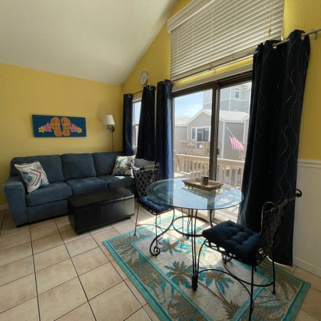 Living room with yellow walls, blue sofa, glass dining table, and large window facing a deck with an American flag.