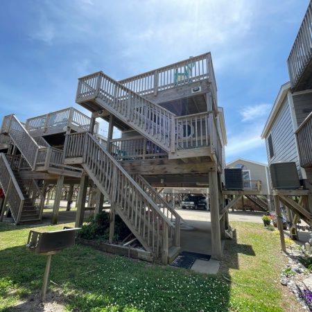 Two-story wooden beach house on stilts with decks, exterior stairs, and a grill in the grassy yard under a blue sky.