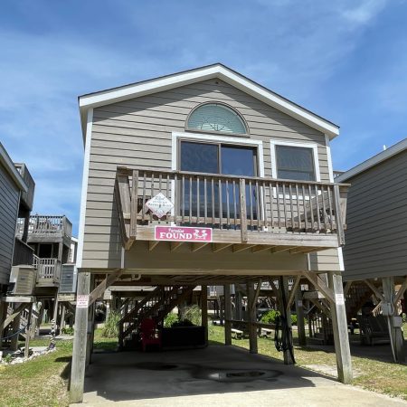 Elevated beach house with wooden deck and “Found” sign, supported by stilts, set against a blue sky.