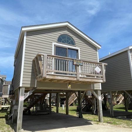 Gray beach house elevated on wooden stilts, with a small front balcony, under a blue sky.
