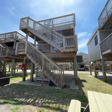 Wooden stilt house with two levels of stairs, a grill, and a picnic table in the foreground on a sunny day.