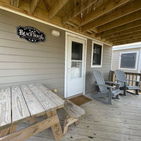 Wooden porch with two chairs, a picnic table, and a door beneath a “Beach House” sign on a beige-sided building.