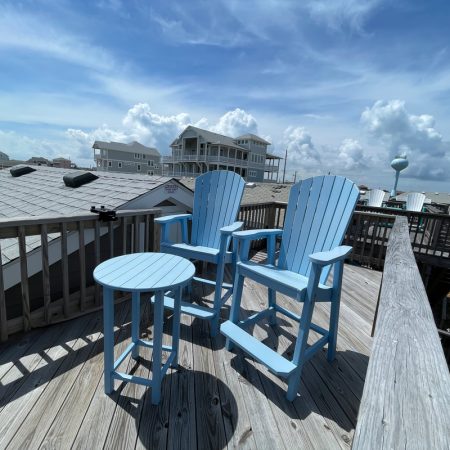 Two blue Adirondack chairs and a round table on a wooden deck overlook houses under a partly cloudy sky.