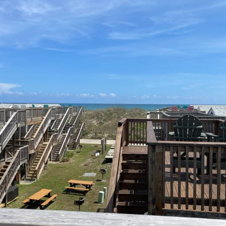 Wooden decks and stairs overlook picnic tables and grassy area, with sand dunes and ocean visible under a blue sky.