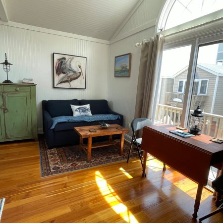 Sunlit living room with a blue couch, wooden table, green cabinet, and a desk near sliding glass doors to a deck.