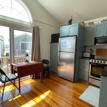Small kitchen with light blue cabinets, stainless appliances, and a desk by a window facing a neighboring house outside.