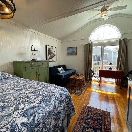 Bright bedroom with a patterned bedspread, sofa, desk, and large window doors leading to a wooden deck outside.