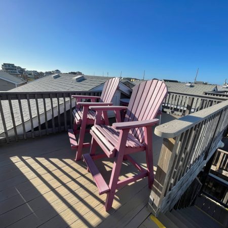 Two pink wooden chairs sit on a rooftop deck overlooking nearby houses under a clear blue sky.