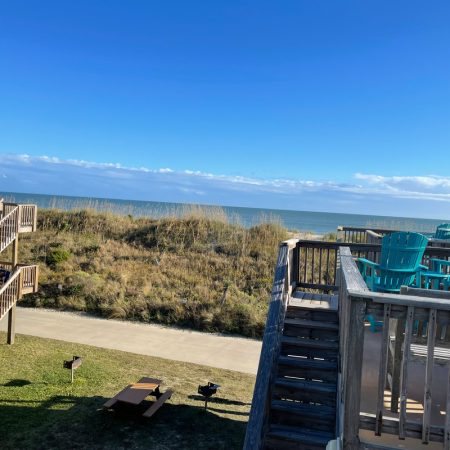 View of a wooden deck with turquoise chairs overlooking sand dunes and the ocean under a clear blue sky.