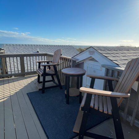 Two wooden chairs and a small table on a rooftop deck with sunlight and a clear sky in the background.