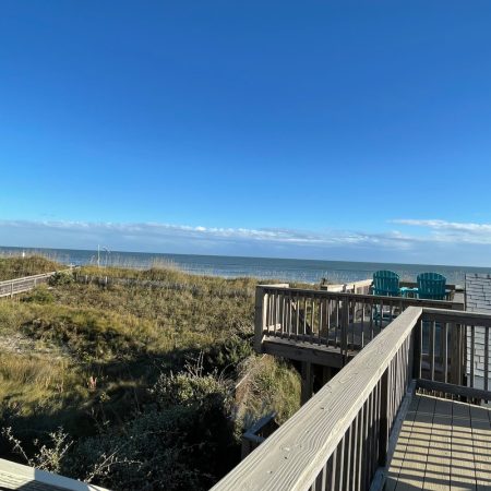 Wooden deck overlooking grassy dunes, two Adirondack chairs, and a clear view of the ocean under a blue sky.