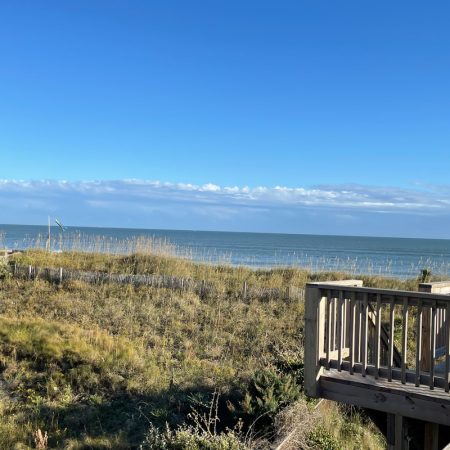 Wooden deck overlooks grassy dunes and the ocean under a clear blue sky with light clouds on the horizon.