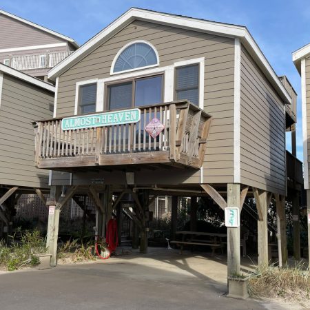 Tan beach house on stilts with a sign reading “Almost Heaven” on the balcony, surrounded by plants and signs.