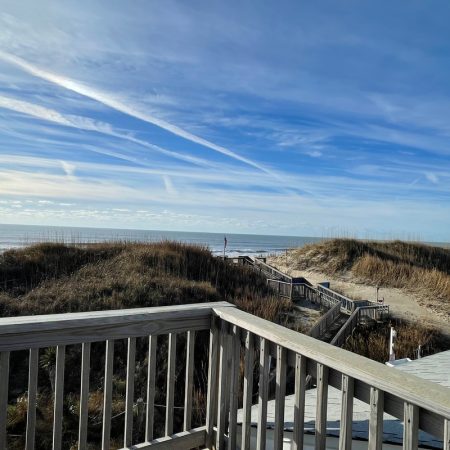 Wooden deck overlooking sand dunes, grass, and a path leading to a beach under a blue sky with wispy clouds.