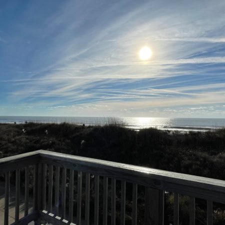 View of the sun over the ocean, seen from a wooden deck with grassy dunes in the foreground.