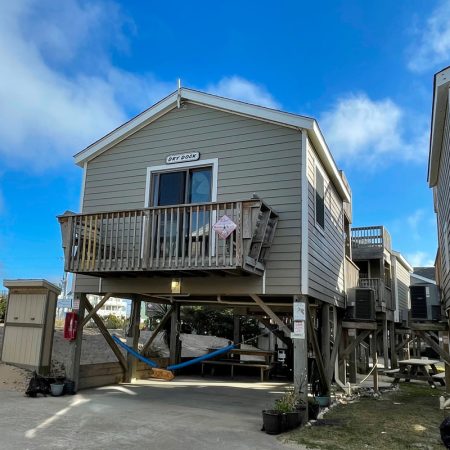 Elevated beach house with a small balcony, hammock below, and a sign on the railing, under a clear blue sky.