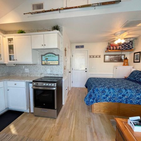 Small studio with white kitchen, bed with blue bedding, ceiling fan, and beach-themed decorations on walls and shelves.