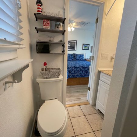 Small bathroom with a toilet, shelves above, and a view into a bedroom with a blue-patterned bedspread.