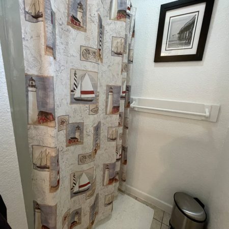 Shower with a nautical-themed curtain, a framed photo on the wall, a towel rack, and a small trash can nearby.