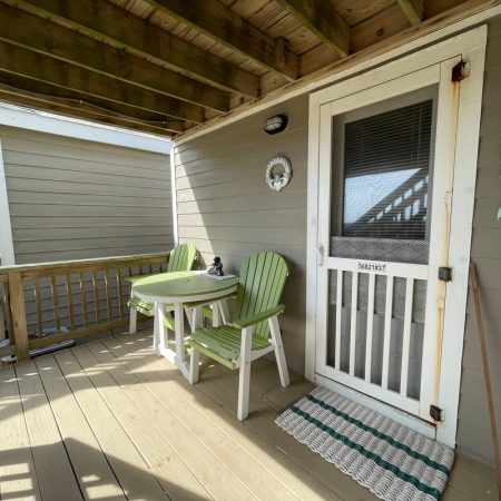 Small porch with two green chairs, a white table, and a screened door labeled “BEACHFRONT” under a wooden overhang.