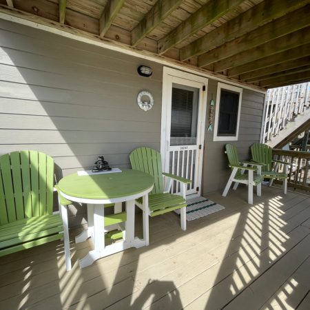 Small porch with green chairs, a round table, wooden railing, and a door under a shaded balcony.