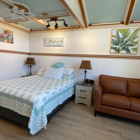 A neatly made bed next to a brown leather loveseat, nightstand, and two lamps in a bright, tidy room.