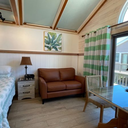 Bright room with a bed, brown loveseat, glass table, chair, lamps, art on walls, and window with green striped curtains.
