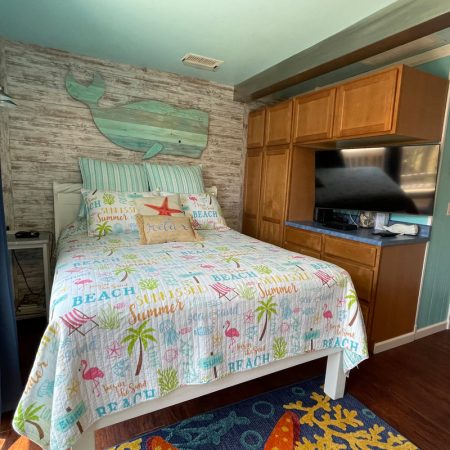 Beach-themed bedroom with a queen bed, wooden whale wall decor, TV, cabinets, and colorful bedding.