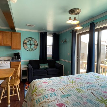 A small beach-themed room with a bed, sofa, kitchen area, and sliding glass door leading to a balcony.
