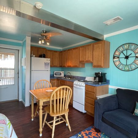 Small kitchen with wood cabinets, dining table, chairs, appliances, blue walls, wall clock, and a navy sofa.