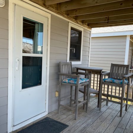 Wood deck with a white door, two tall chairs, a table, and a window on the gray siding wall outside a house.