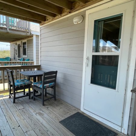 Small wooden deck with a table, chairs, and a bench next to a white door on a gray house.