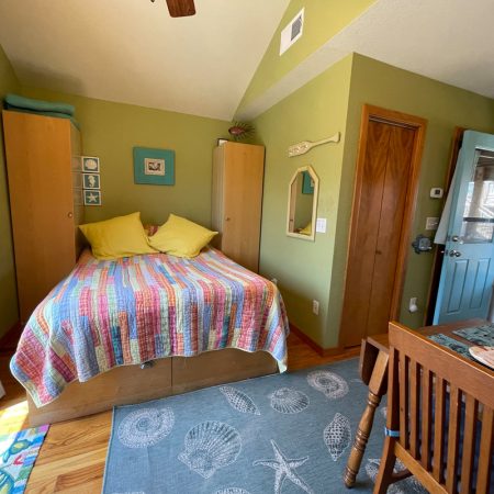 Small bedroom with a bed, colorful quilt, yellow pillows, wooden furniture, green walls, and a blue door.