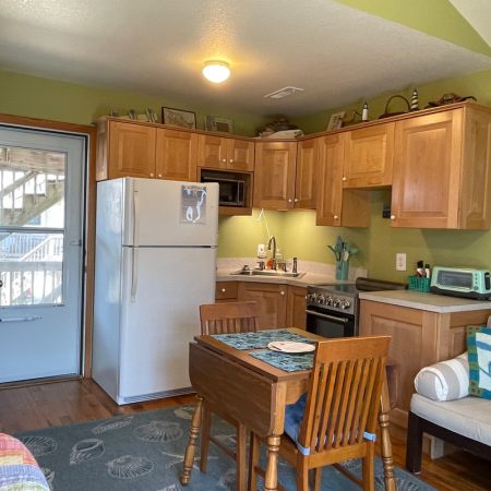 Small kitchen with wooden cabinets, dining table for two, white fridge, stove, and a cozy seating area with a pillow.
