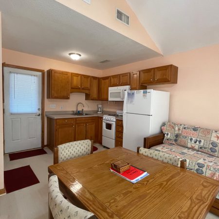 Small kitchen and living area with wooden cabinets, table, chairs, patterned couch, and a refrigerator.
