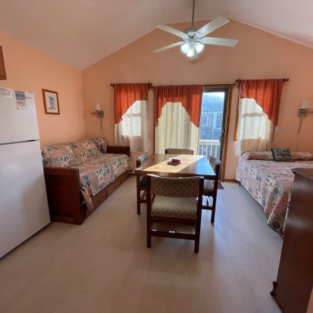 Small room with a fridge, two beds, a table with chairs, ceiling fan, and sunlight coming through curtained windows.