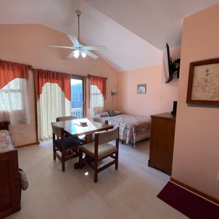 A cozy bedroom with peach walls, a bed, a dining table, chairs, a dresser, and windows with red curtains.