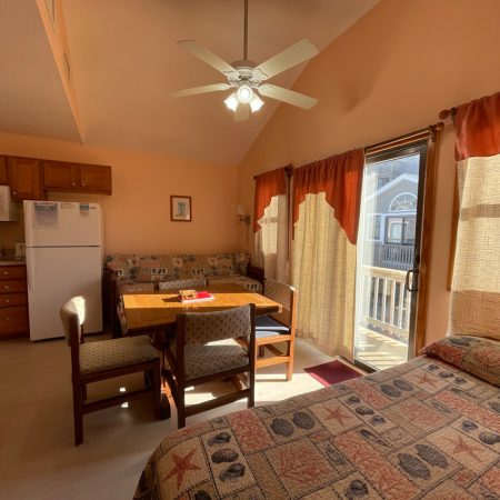 Sunny studio apartment with bed, table, kitchen area, and sliding glass doors leading to a balcony.