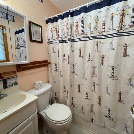 Small bathroom with a white sink, toilet, and a lighthouse-themed shower curtain. Mirror and wall art above sink.