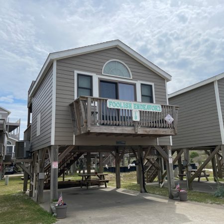 Elevated gray beach house with a small balcony and sign reading “Foolish Endeavor” above the door, under a cloudy sky.
