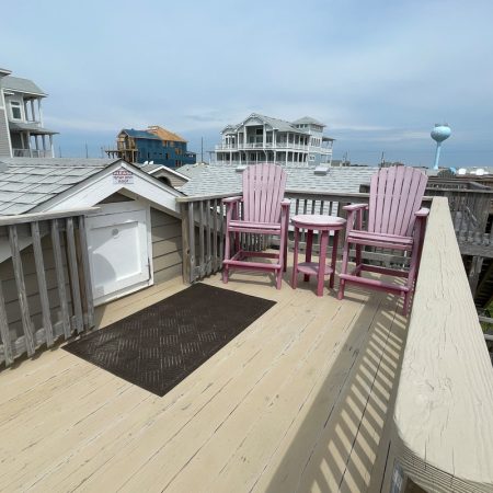 Two pink wooden chairs and a small table on a rooftop deck overlooking neighboring beach houses on a cloudy day.