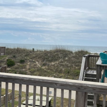 Wooden deck overlooks grassy dunes and ocean under a cloudy sky, with blue chairs placed on the decks.