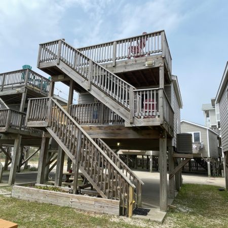 Two-story stilt beach house with wooden stairs and railings, elevated above ground with parking space underneath.