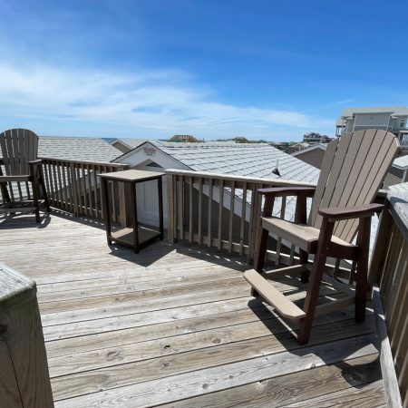 Wooden deck with two chairs and two small tables, overlooking rooftops and a blue sky on a sunny day.