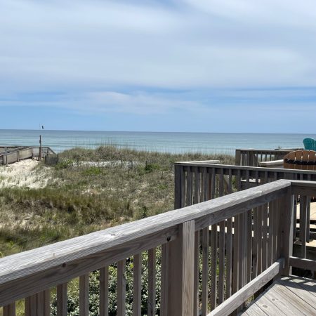 Wooden deck overlooks grassy dunes and calm ocean under a partly cloudy sky, with chairs visible on the deck.