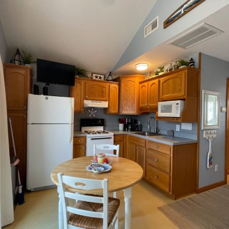 Small kitchen with wooden cabinets, white appliances, round dining table, and two chairs in a cozy open living space.